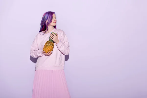 Beautiful young woman with colorful hair and pink sweatshirt holding pineapple on pink — Stock Photo