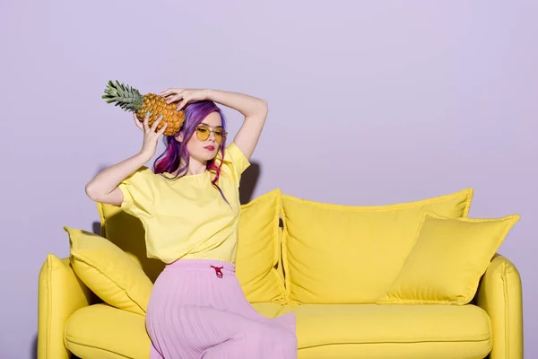 Красива молода жінка сидить на жовтому дивані і тримає ананас біля голови — стокове фото