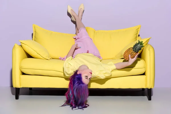 Beautiful young woman lying upside down on yellow couch with pineapple — Stock Photo