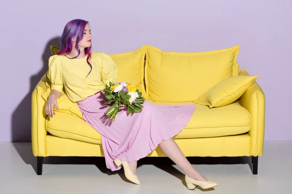 Beautiful young woman sitting on yellow couch with floral bouquet — Stock Photo