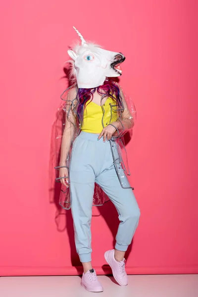 Donna elegante in impermeabile trasparente alla moda e maschera di unicorno su rosso — Foto stock