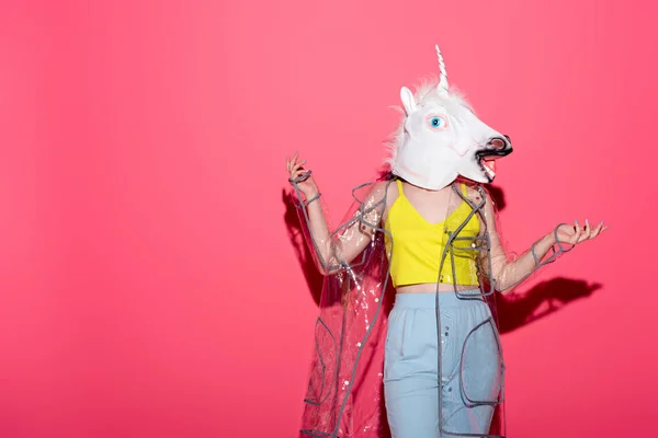 Funny woman in fashionable transparent raincoat and unicorn mask on red — Stock Photo