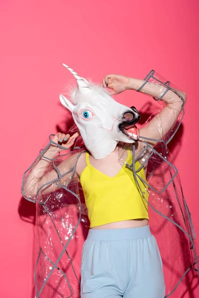 Ridiculous woman in fashionable transparent raincoat and unicorn mask on red — Stock Photo