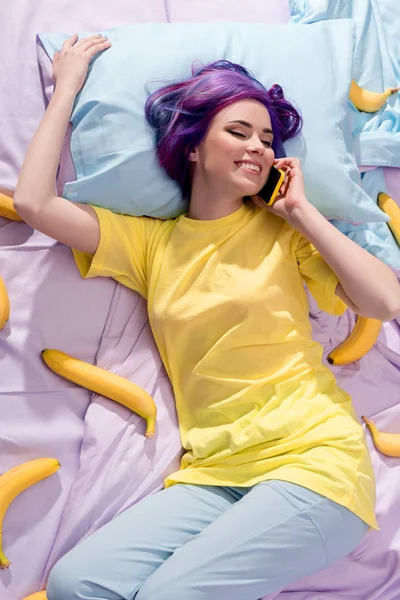 Visão de alto ângulo de feliz jovem deitada na cama com bananas e falando por telefone — Fotografia de Stock