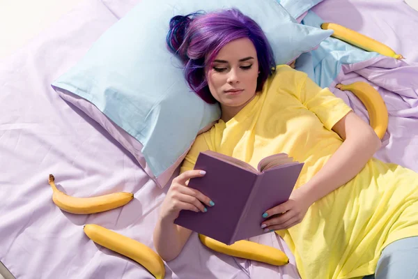 Vista ad alto angolo di giovane donna sdraiata a letto con banane e libro di lettura — Foto stock