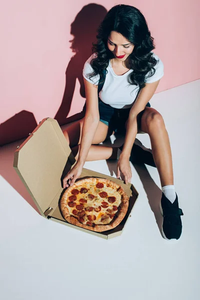 Vue grand angle de la femme élégante et boîte de livraison avec pizza sur le sol sur fond rose — Photo de stock