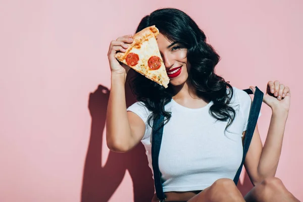 Vista oscurecida de hermosa mujer sonriente que cubre el ojo con un pedazo de pizza sobre fondo rosa - foto de stock