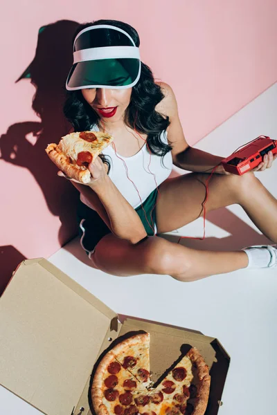 High angle view of stylish woman in cap with retro music player holding piece of pizza on pink backdrop — Stock Photo