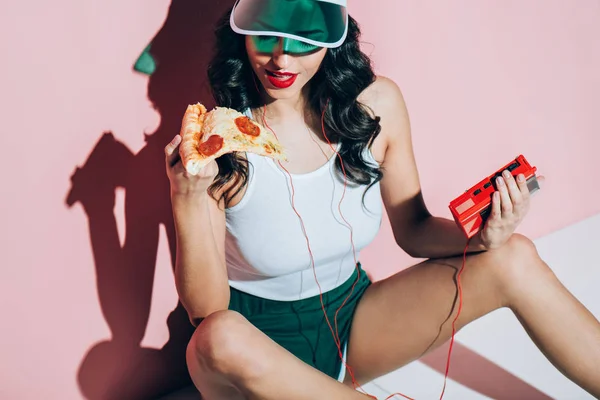 Femme élégante en casquette avec lecteur de musique rétro tenant morceau de pizza sur fond rose — Photo de stock