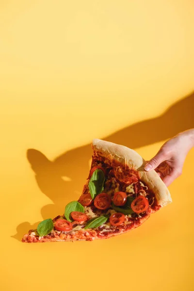 Vista parcial de la mujer sosteniendo un pedazo de pizza con tomates cherry en la mano sobre fondo amarillo - foto de stock