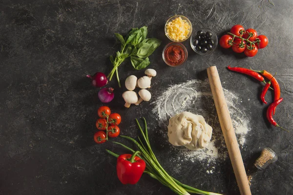 Flat lay with rolling pin, raw dough and fresh ingredients for pizza on dark surface — Stock Photo