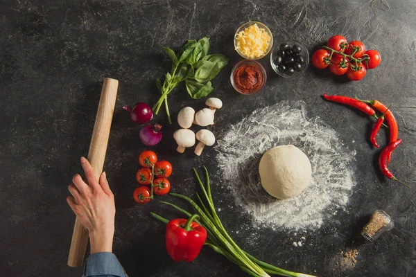 Tiro recortado de mão feminina, ingredientes frescos e massa crua para pizza italiana em mesa escura — Fotografia de Stock