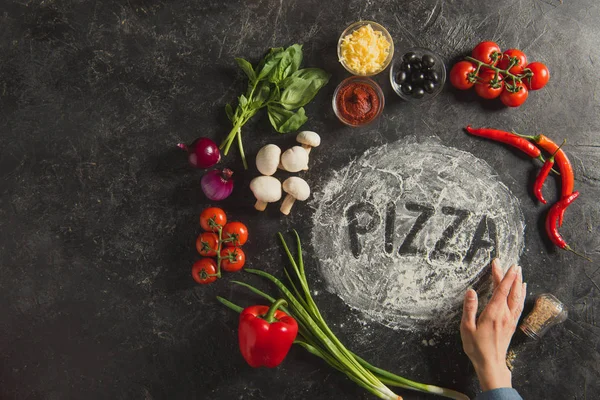 Partial view of female hand, fresh ingredients and pizza lettering made of flour on dark surface — Stock Photo