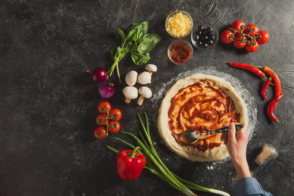 Cropped shot of woman putting sauce on raw dough while cooking italian pizza on dark tabletop — Stock Photo