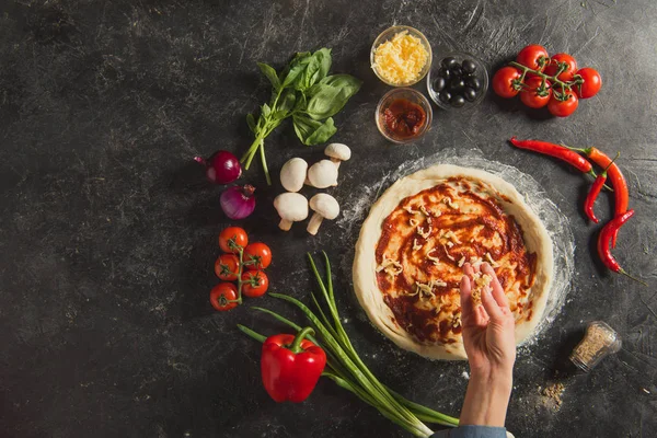Обрезанный снимок женщины, кладущей тертый сыр на тесто во время приготовления итальянской пиццы на темном столе — стоковое фото