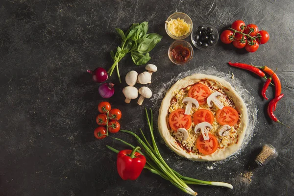 Top view of raw italian pizza and fresh ingredients around on dark surface — Stock Photo