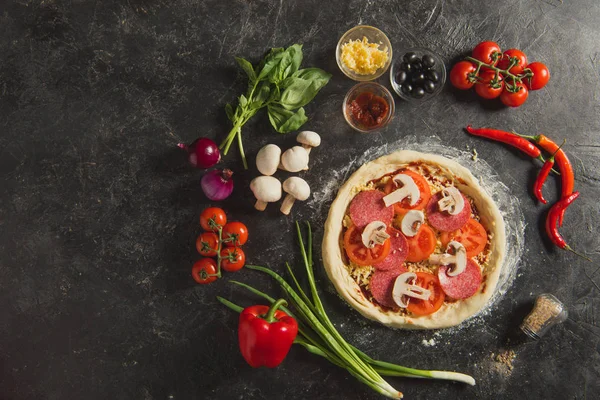 Top view of raw italian pizza and fresh ingredients around on dark surface — Stock Photo