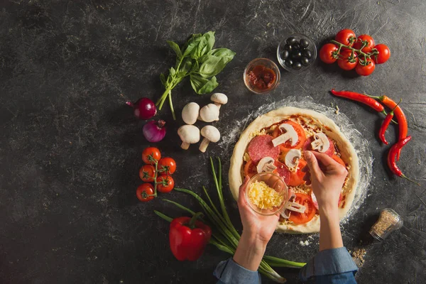 Tiro recortado de la mujer poniendo queso rallado en la masa mientras cocina pizza italiana en la mesa oscura - foto de stock