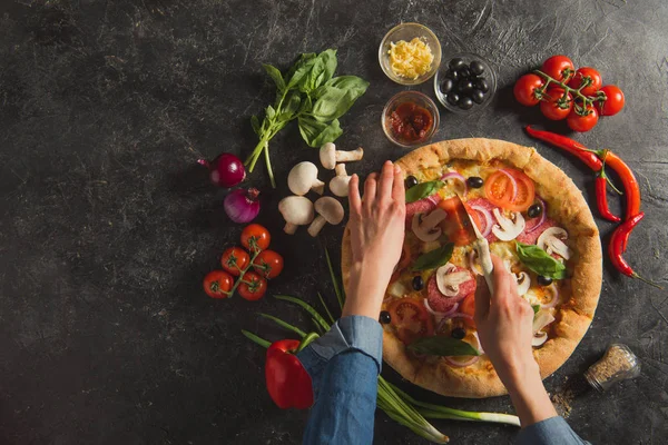 Обрезанный снимок женщины, резающей приготовленную итальянскую пиццу со свежими ингредиентами на темной столешнице — стоковое фото