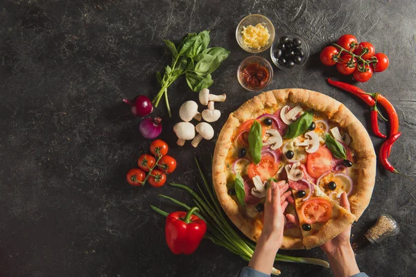 Cropped shot of female hands and cooked italian pizza with fresh ingredients on dark tabletop — Stock Photo