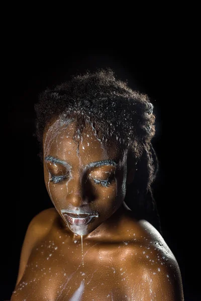 Соблазнительная африканская девушка с молочными капельками на лице и теле, позирующая с закрытыми глазами, изолированными на черном — стоковое фото