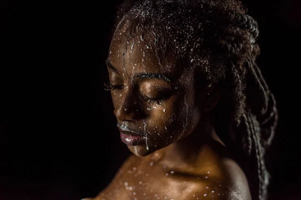 Primo piano vista di seducente ragazza afroamericana con gocce di latte sul viso e sul corpo isolato su nero — Foto stock