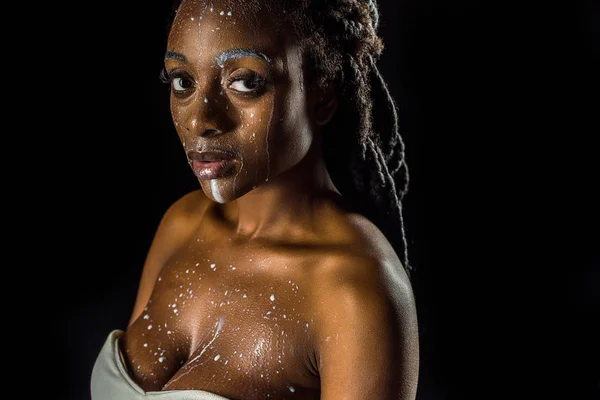 Sensuelle jeune afro-américaine avec du lait renversé sur le visage en regardant la caméra isolée sur noir — Photo de stock