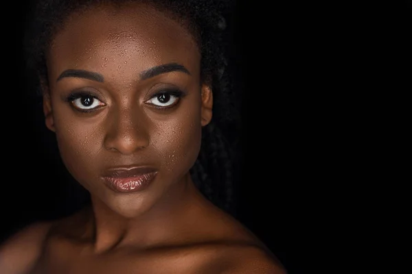 Porträt der schönen jungen afrikanisch-amerikanischen Frau mit Wassertropfen im Gesicht, die in die Kamera schaut, isoliert auf schwarz — Stockfoto