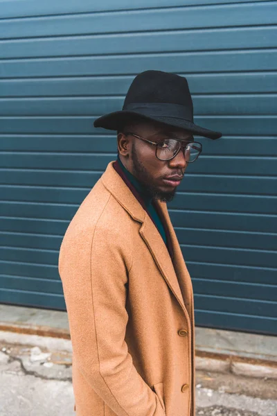 Portrait of stylish young african american man in hat, eyeglasses and overcoat looking at camera — Stock Photo