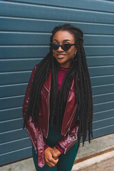 Hermosa sonriente joven afroamericana mujer en chaqueta y gafas de sol posando en la calle - foto de stock