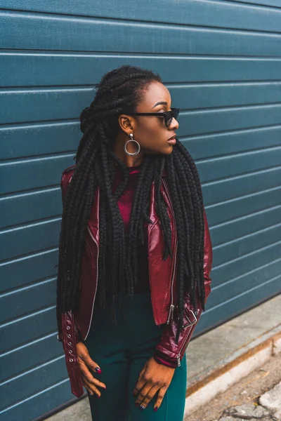 Bela jovem mulher americana africana elegante em jaqueta e óculos de sol olhando para longe na rua — Fotografia de Stock