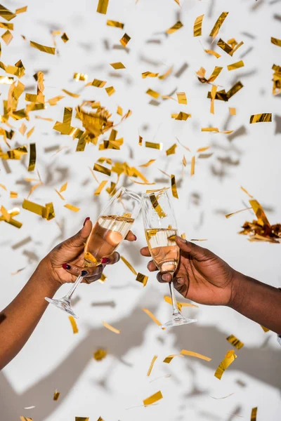 Cropped shot of african american couple clinking glasses of champagne on white with golden confetti — Stock Photo