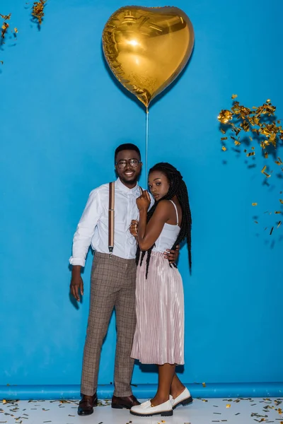 Elegante joven africano americano pareja posando con oro corazón en forma de globo en azul - foto de stock