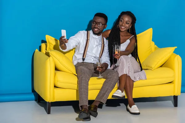 Sorrindo jovem casal afro-americano segurando copos de vinho e tomando selfie com smartphone no sofá — Fotografia de Stock