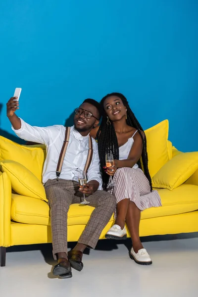 Glücklich stylisches junges afrikanisch-amerikanisches Paar hält Weingläser in der Hand und macht Selfie mit Smartphone auf dem Sofa — Stockfoto