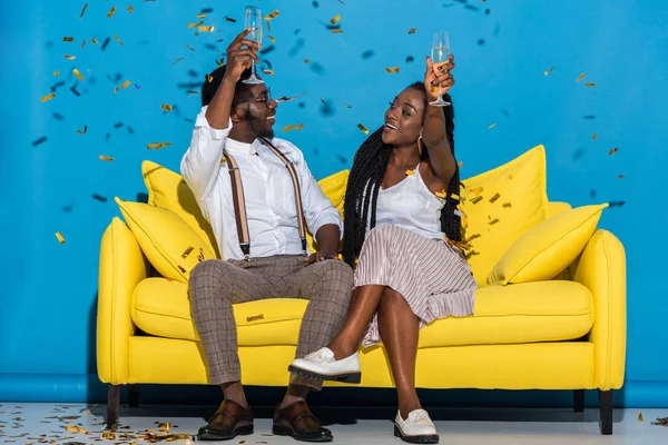 Alegre elegante joven africano americano pareja sosteniendo vasos de vino y sonriendo entre sí mientras se sienta en el sofá bajo la caída de confeti - foto de stock