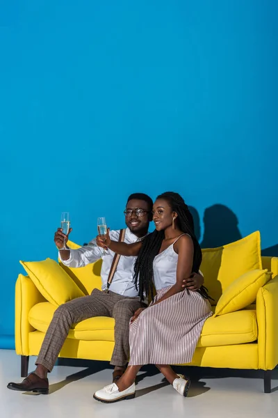 Alegre elegante jovem casal afro-americano segurando copos de vinho enquanto sentado no sofá — Fotografia de Stock