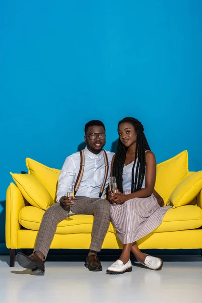 Joven pareja afroamericana sosteniendo copas de vino y sonriendo a la cámara mientras se sienta en un sofá amarillo - foto de stock