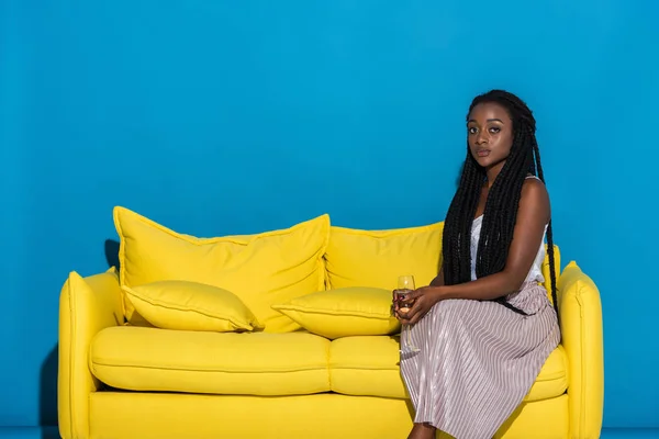 Bela jovem afro-americana segurando copo de vinho enquanto sentado no sofá e olhando para a câmera em azul — Fotografia de Stock