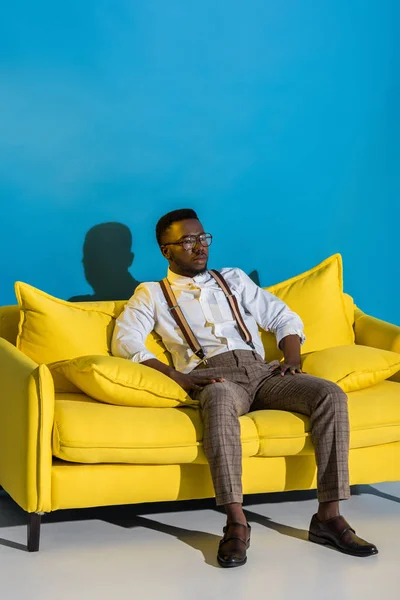 Stylischer junger afrikanisch-amerikanischer Mann mit Brille sitzt auf gelbem Sofa und schaut weg auf blauem Grund — Stockfoto