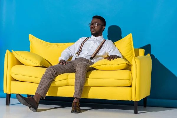 Beau jeune homme afro-américain élégant assis sur un canapé jaune et regardant loin sur le bleu — Photo de stock