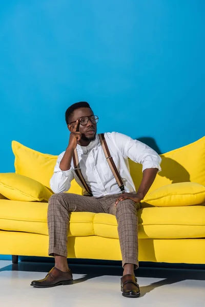 Beau jeune homme afro-américain élégant assis sur le canapé jaune sur le bleu — Photo de stock