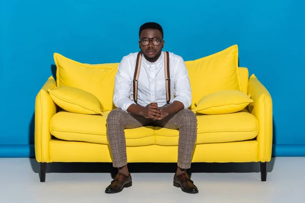 Молодой африканский американец в очках сидит на диване и смотрит в камеру — стоковое фото