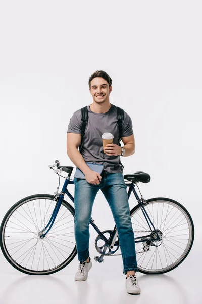 Studente sorridente in possesso di caffè per andare e appoggiato in bicicletta sul bianco — Foto stock