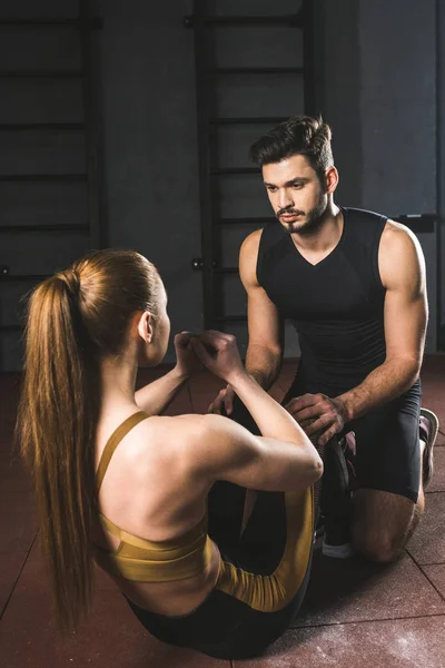 Entrenador masculino ayudando a la joven deportista a hacer abdominales en el gimnasio - foto de stock