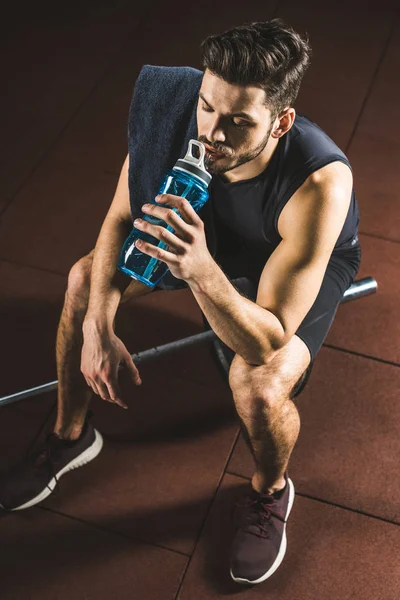 Hochwinkelaufnahme des Sportlers, der Wasser aus der Flasche trinkt und in der Turnhalle auf der Langhantel sitzt — Stockfoto