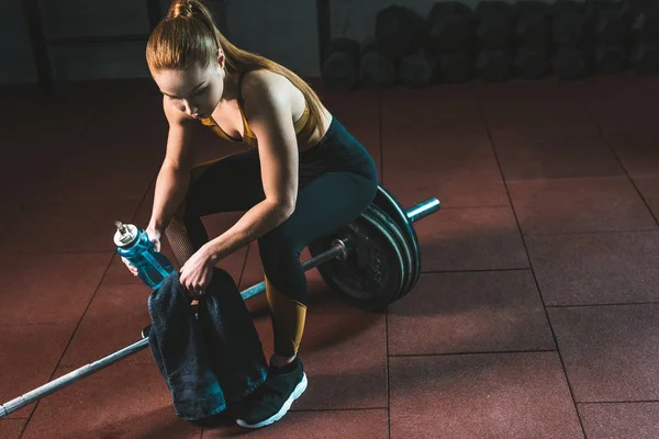 Tired sportswoman with bottle of water and towel sitting on barbell in gym — Stock Photo