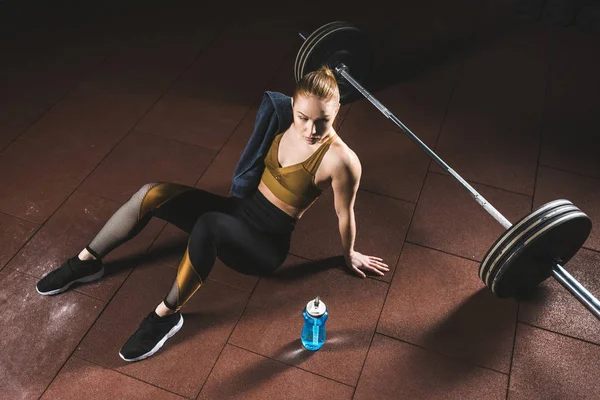 Young sportswoman with towel on shoulder sitting on floor near barbell in gym — Stock Photo