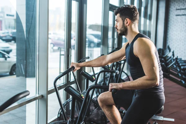 Vista lateral del joven deportista haciendo ejercicio en bicicleta estática en el centro deportivo — Stock Photo