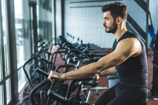 Vista lateral del joven deportista haciendo ejercicio en bicicleta estática en el centro deportivo - foto de stock